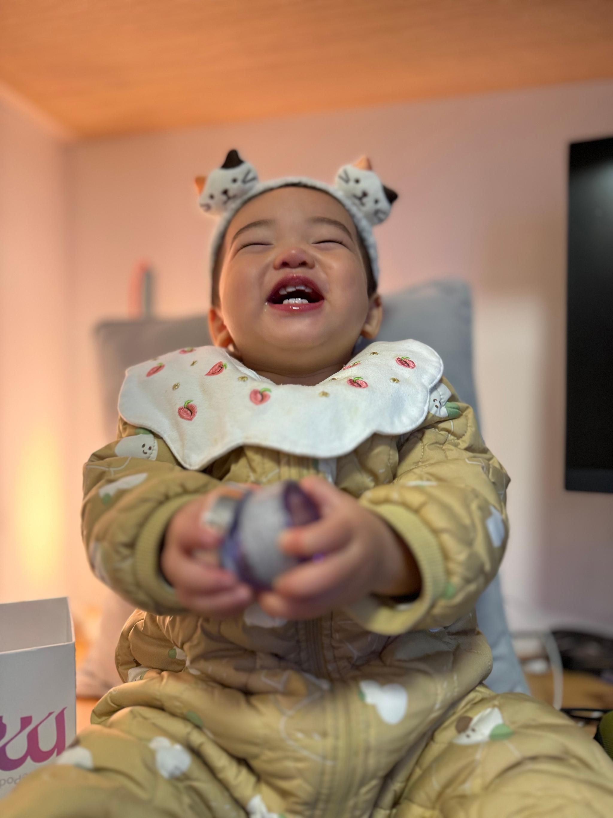 A laughing baby dressed in a yellow outfit with decorative frills and patterns, wearing a headband with cute ears, holding a small ball