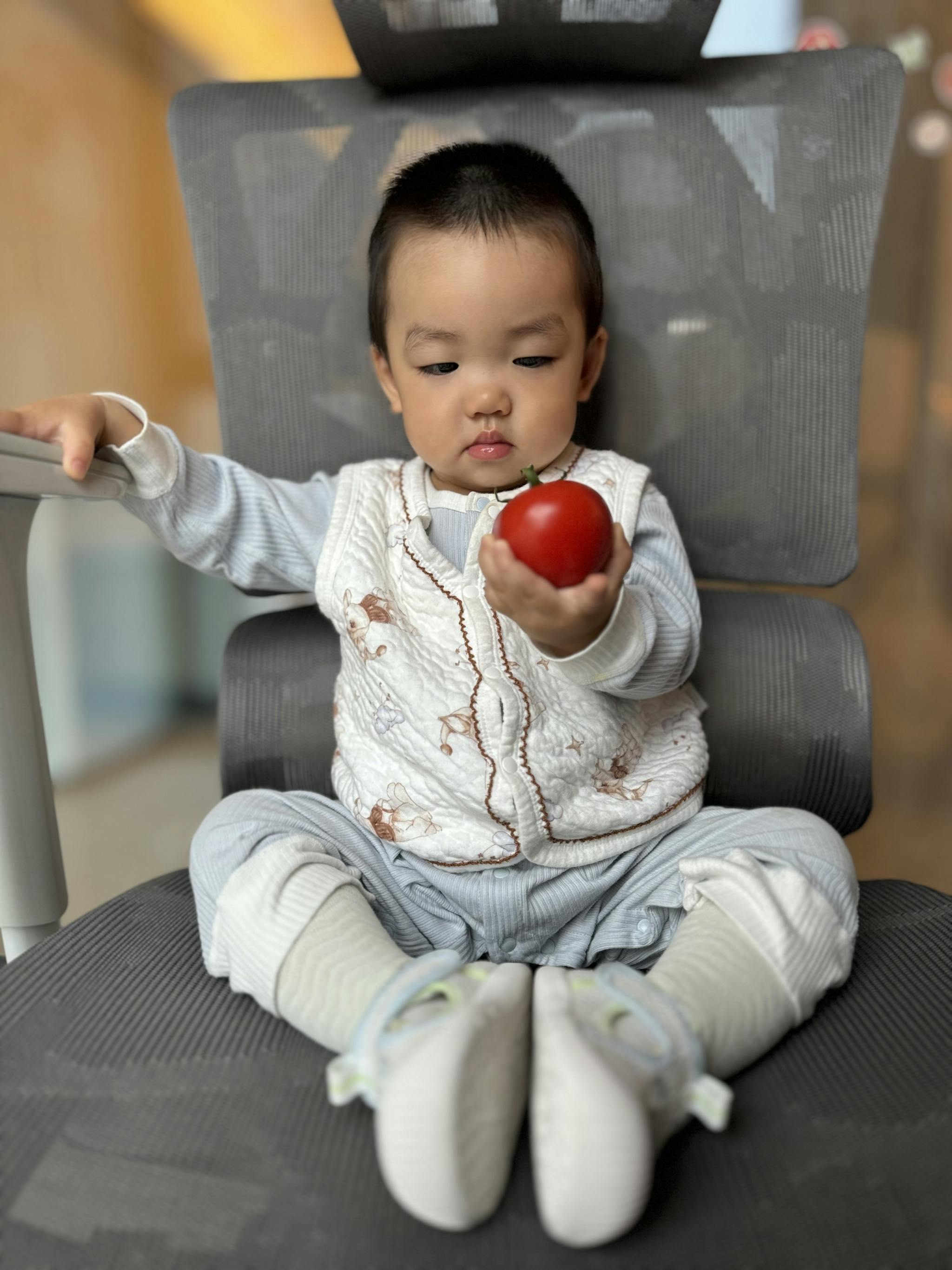 A young child sitting cross-legged on a chair extends their arm holding a red ball, with a focused expression on their face