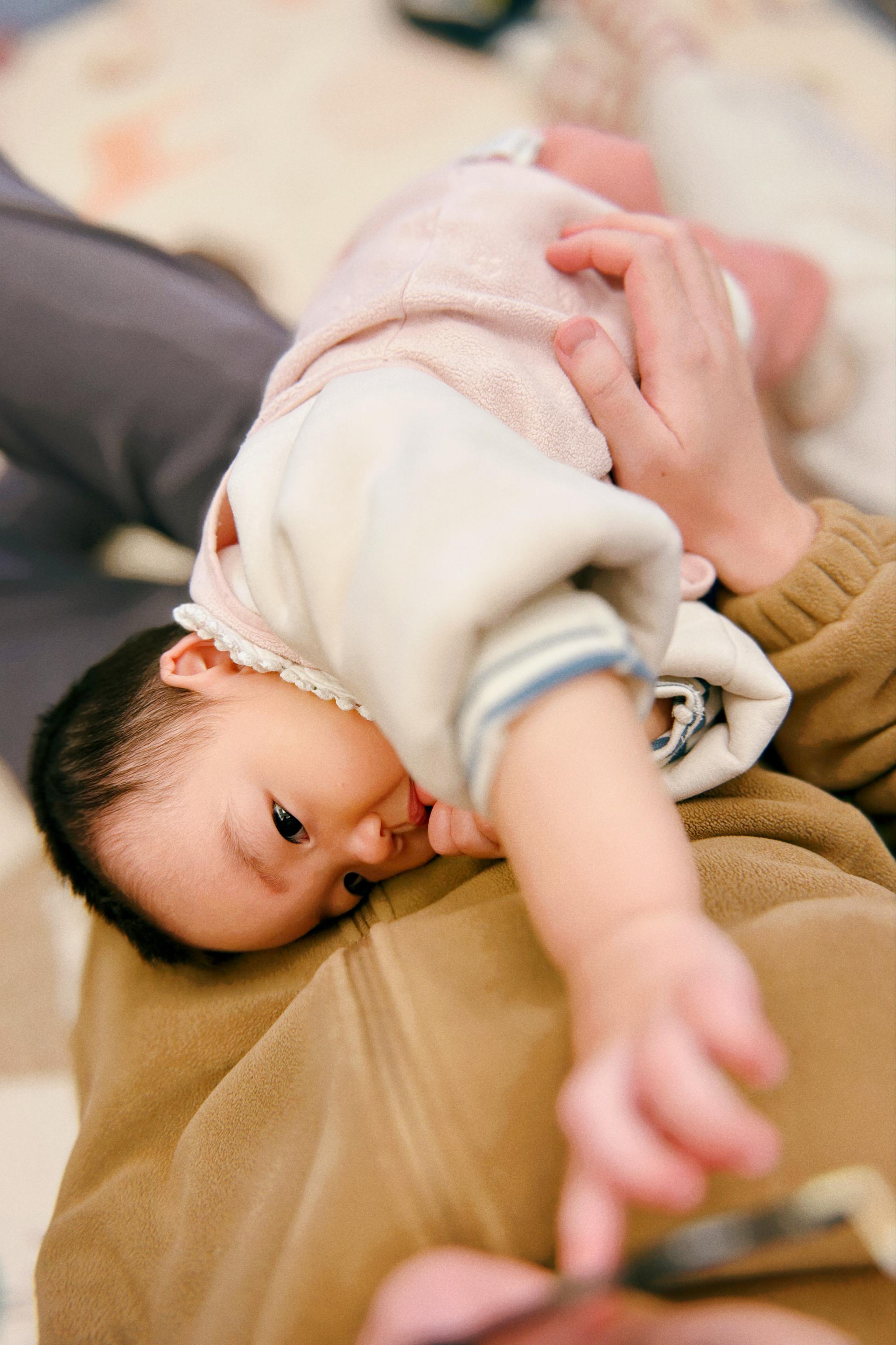 A baby dressed in pink is lying on top of an adult, reaching out towards the camera, with a blurred background emphasizing the child