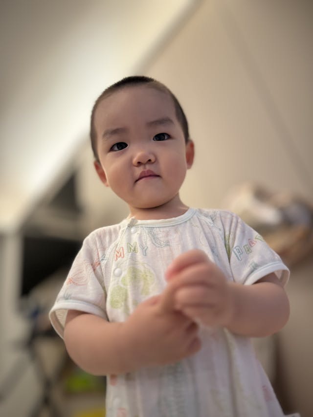 A young child with short hair, wearing a light-colored shirt, stands indoors with a neutral expression, holding their hands together The background is softly blurred