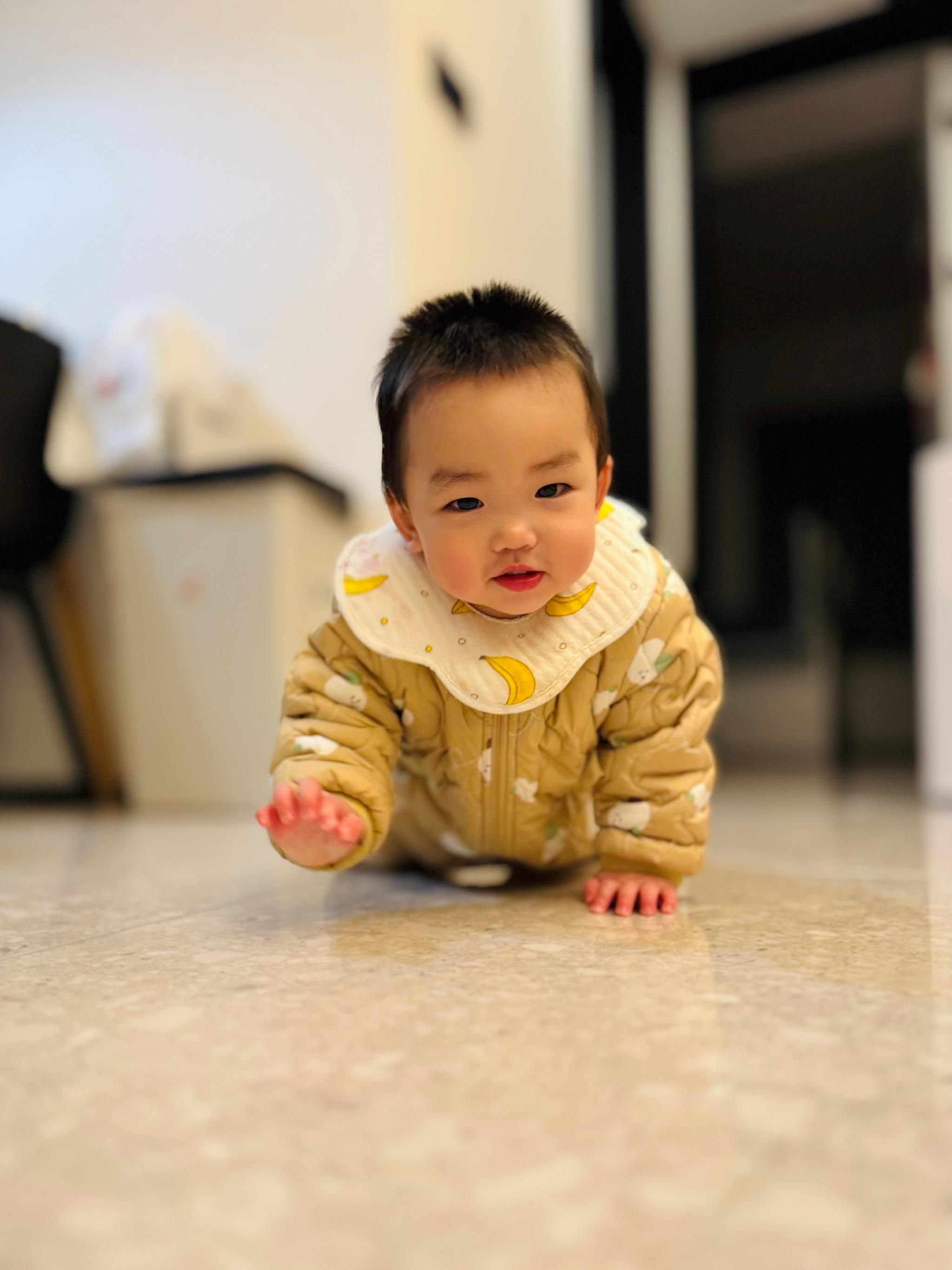 A baby in a yellow outfit is on all fours on a floor, looking forward with a curious expression