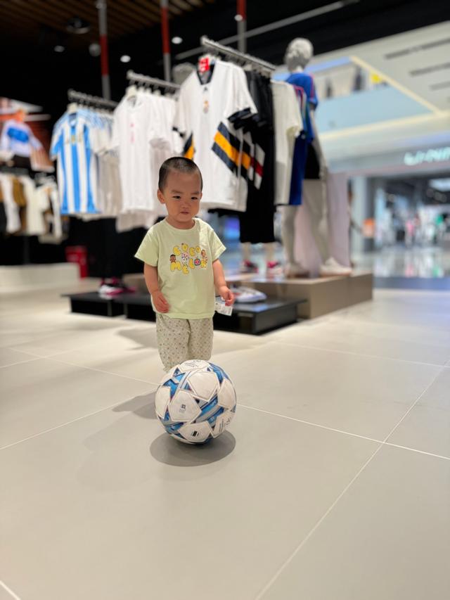 A young child stands in a clothing store, focused on a soccer ball at their feet