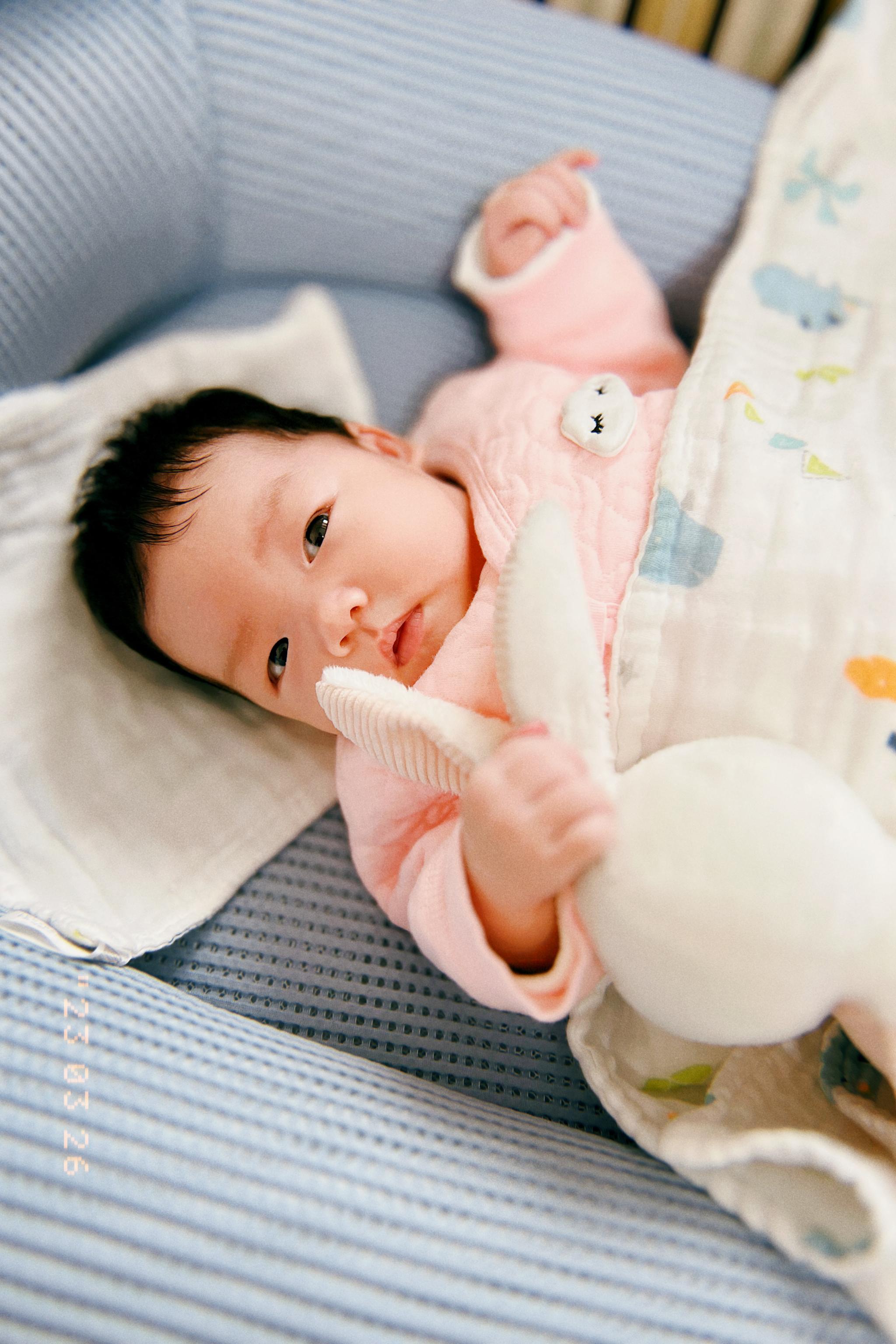 A baby dressed in pink, lying on a blue blanket, looks directly at the camera while holding a white balloon