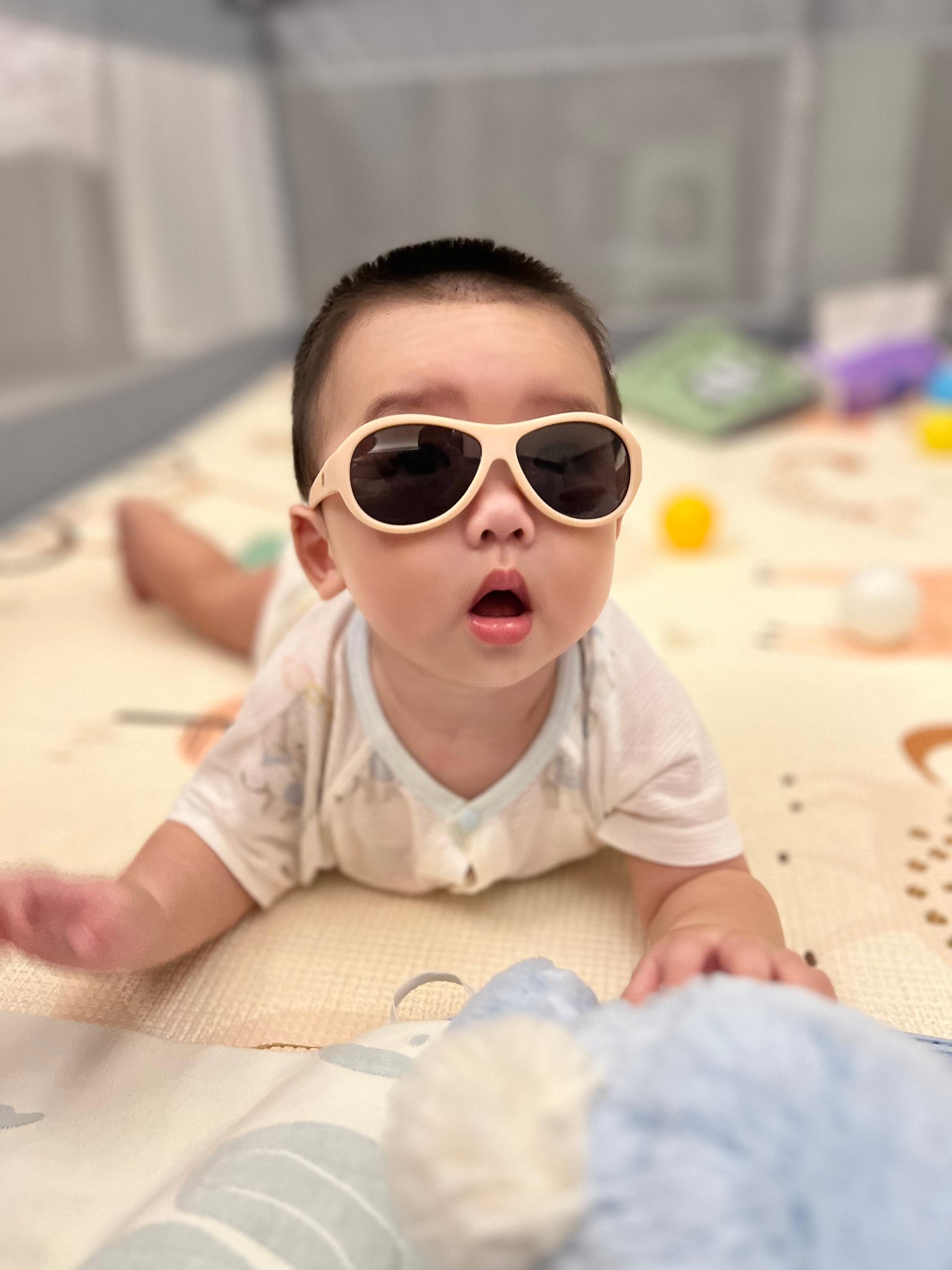 A baby wearing oversized sunglasses lies on a patterned blanket, exhibiting a look of surprise or fascination