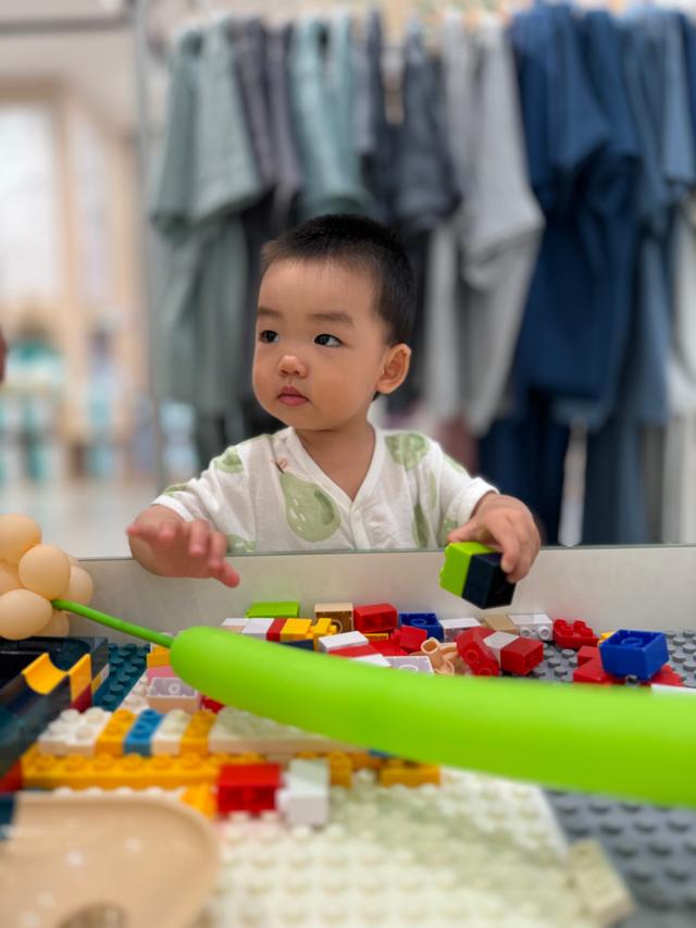 A toddler is focused on playing with colorful blocks on a green table, with a blurred background suggesting a retail environment