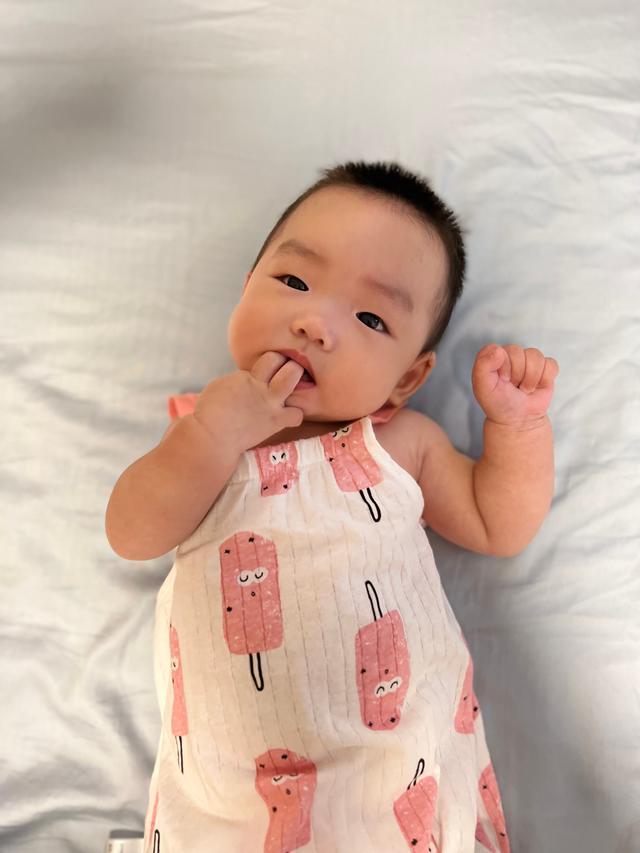A baby with fingers in mouth wearing a sleeveless outfit with pink patterns, lying on a white background