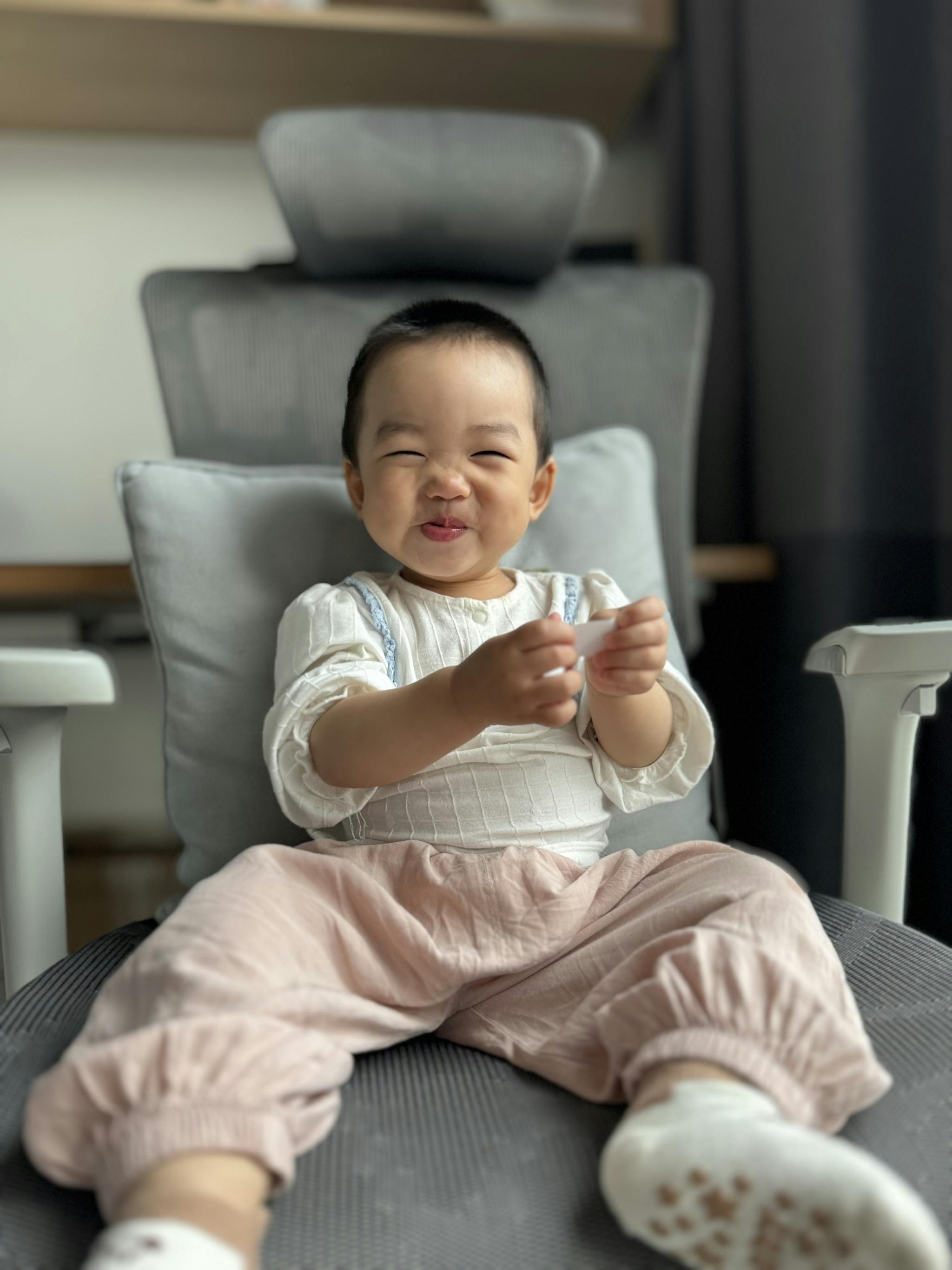 A baby with a humorous expression sits on a cushioned chair, sticking out their tongue and squinting one eye, dressed in a white top and pink pants