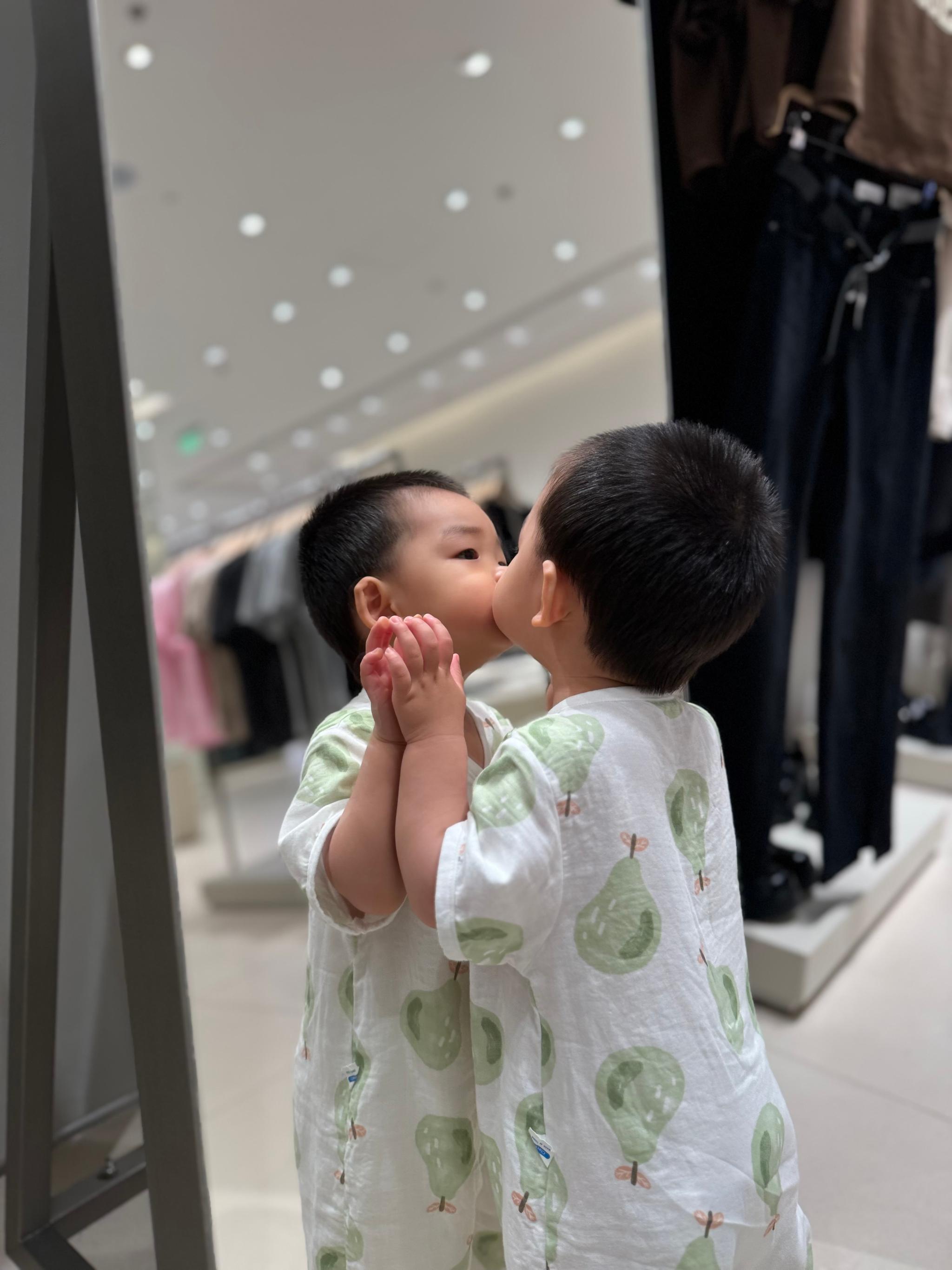 A young child in a green patterned outfit kisses their reflection in a mirror, standing in a clothing store