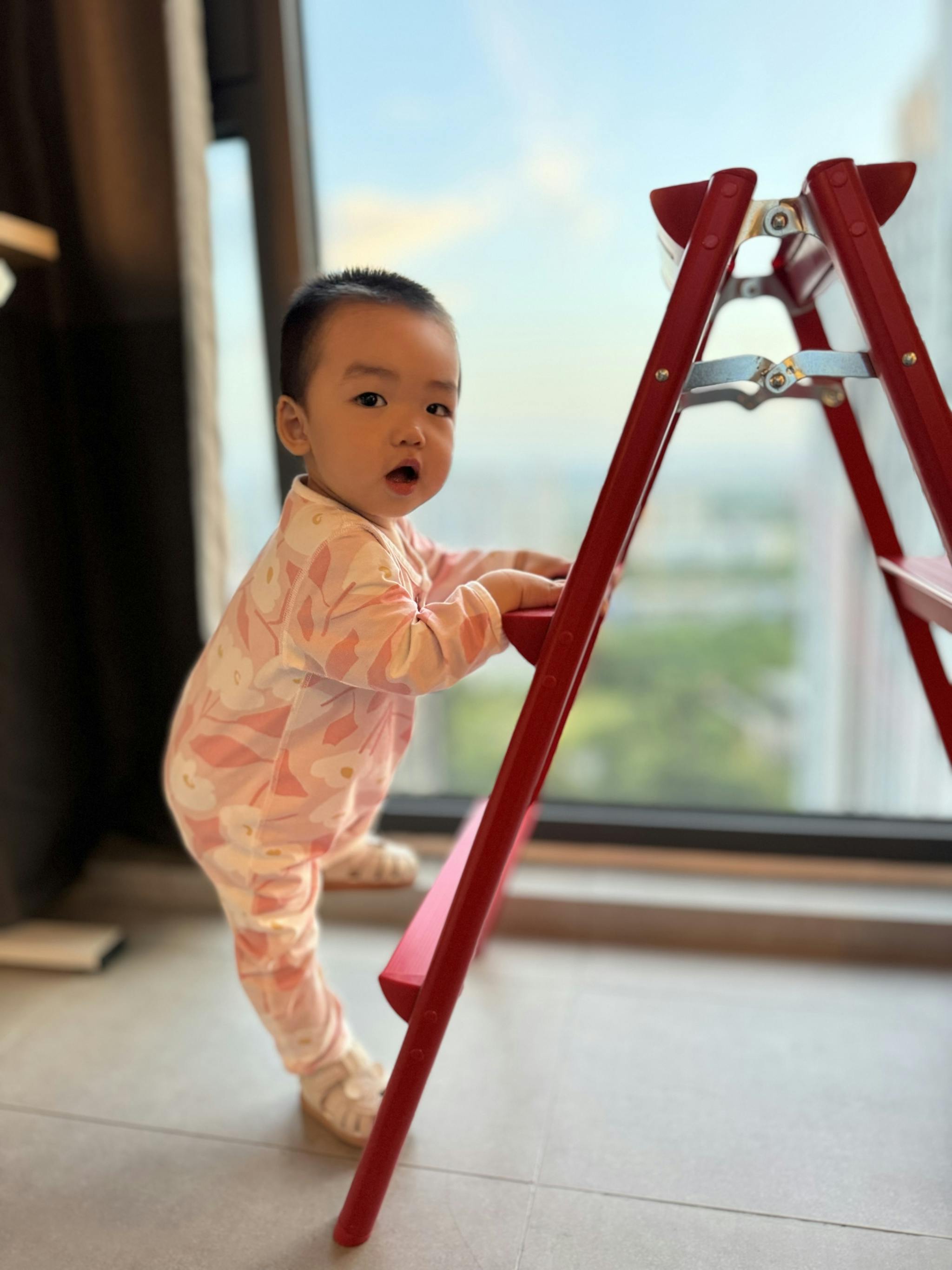 A toddler in pink pajamas stands by a red ladder near a window, looking toward the camera