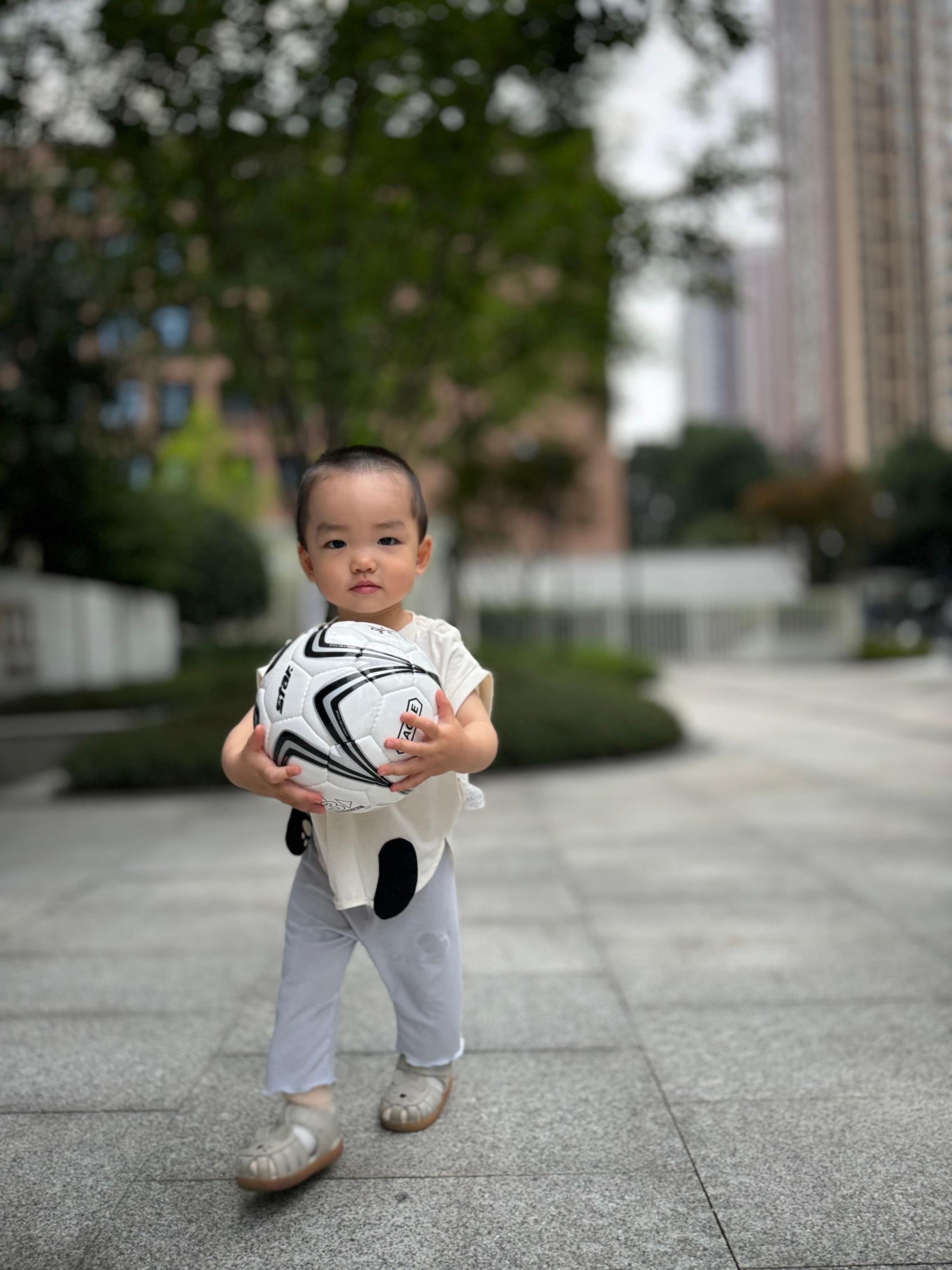 A young child holding a soccer ball stands on a paved area with trees and buildings in the background