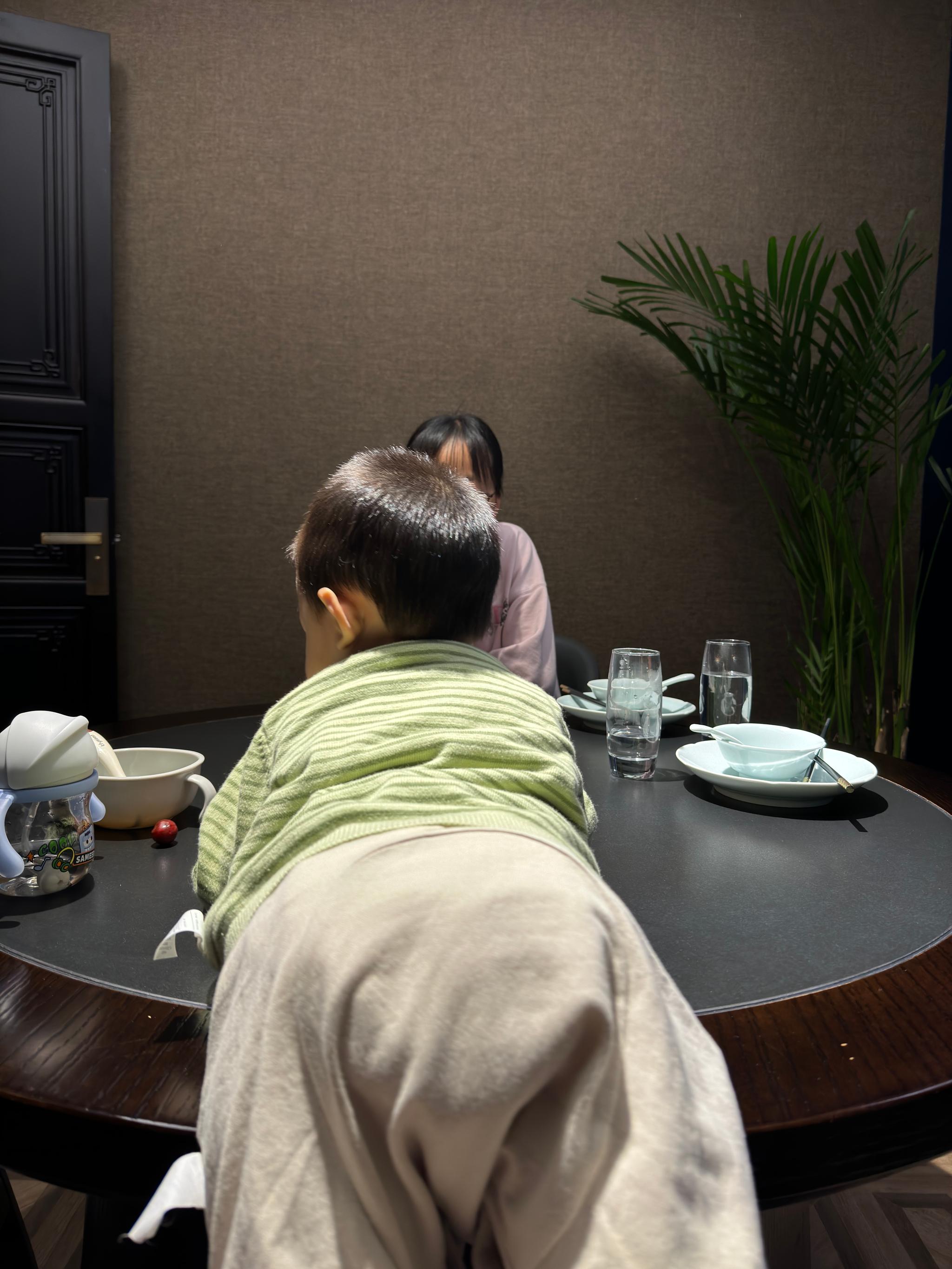 A person seated at a dining table with an empty plate and utensils in front of them, appearing to be in conversation with someone whose back is facing the camera. A plant is visible in the background, suggesting a comfortable indoor setting