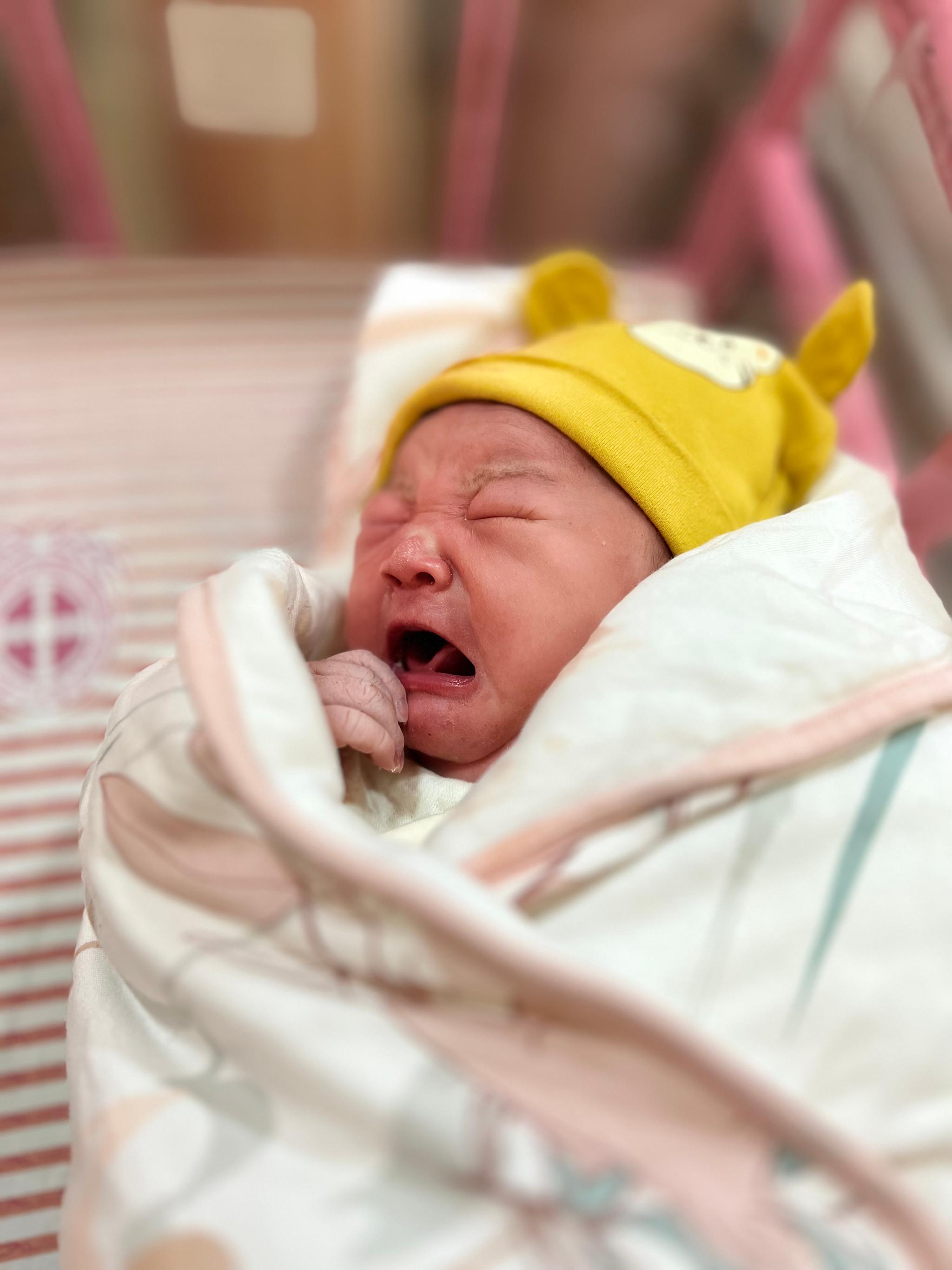 A newborn baby wearing a yellow hat appears to be yawning or crying while wrapped in a blanket