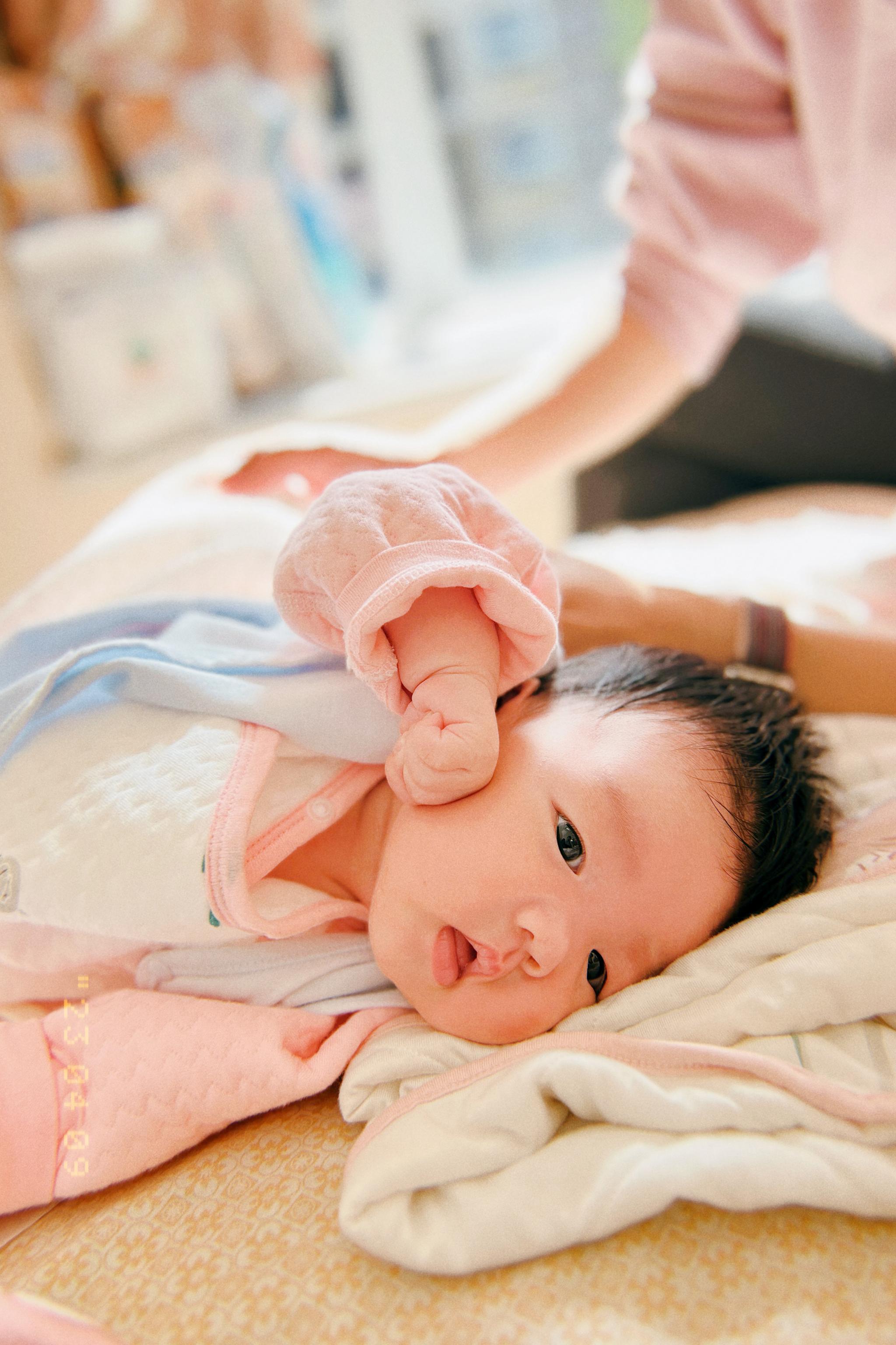 An infant dressed in pink is lying down, looking alert, with an adult's hand gently touching the baby's arm