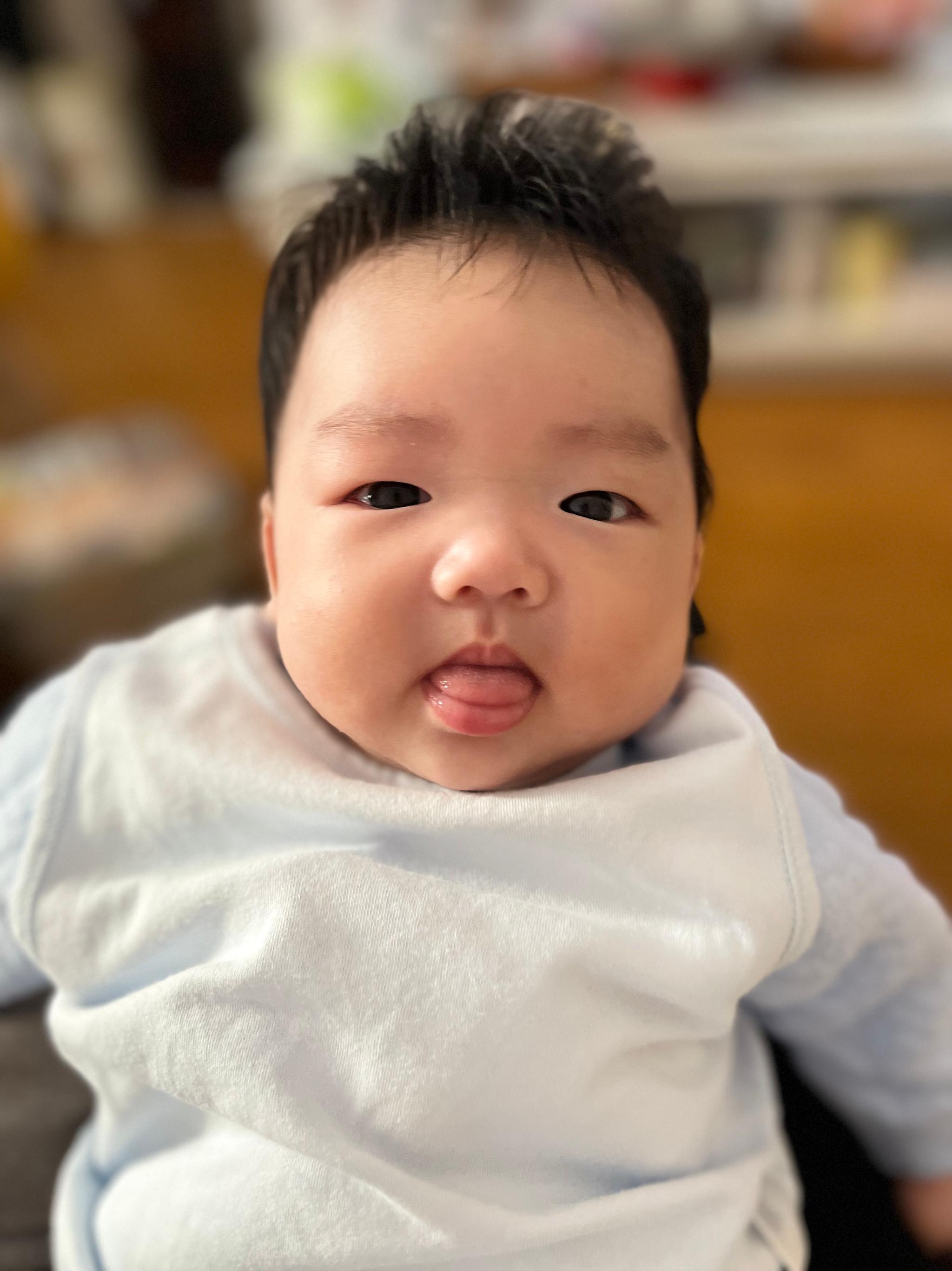 A baby with a tuft of black hair is wrapped in a white cloth, poking its tongue out slightly, and looking directly at the camera