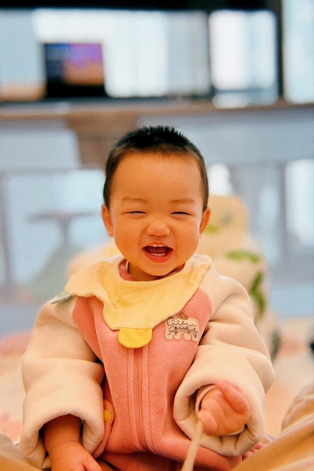 A joyful baby with a big smile, wearing a pink bib over a cozy outfit, with eyes closed in a laugh or giggle