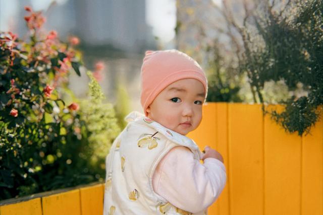 A toddler in a pink hat and patterned outfit looks over their shoulder against a bright yellow fence with flowers in the background