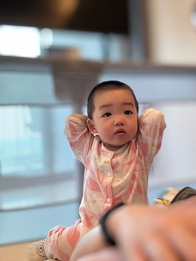 A toddler in a pink outfit with arms stretched upwards, looking slightly to the side, with a blurred background