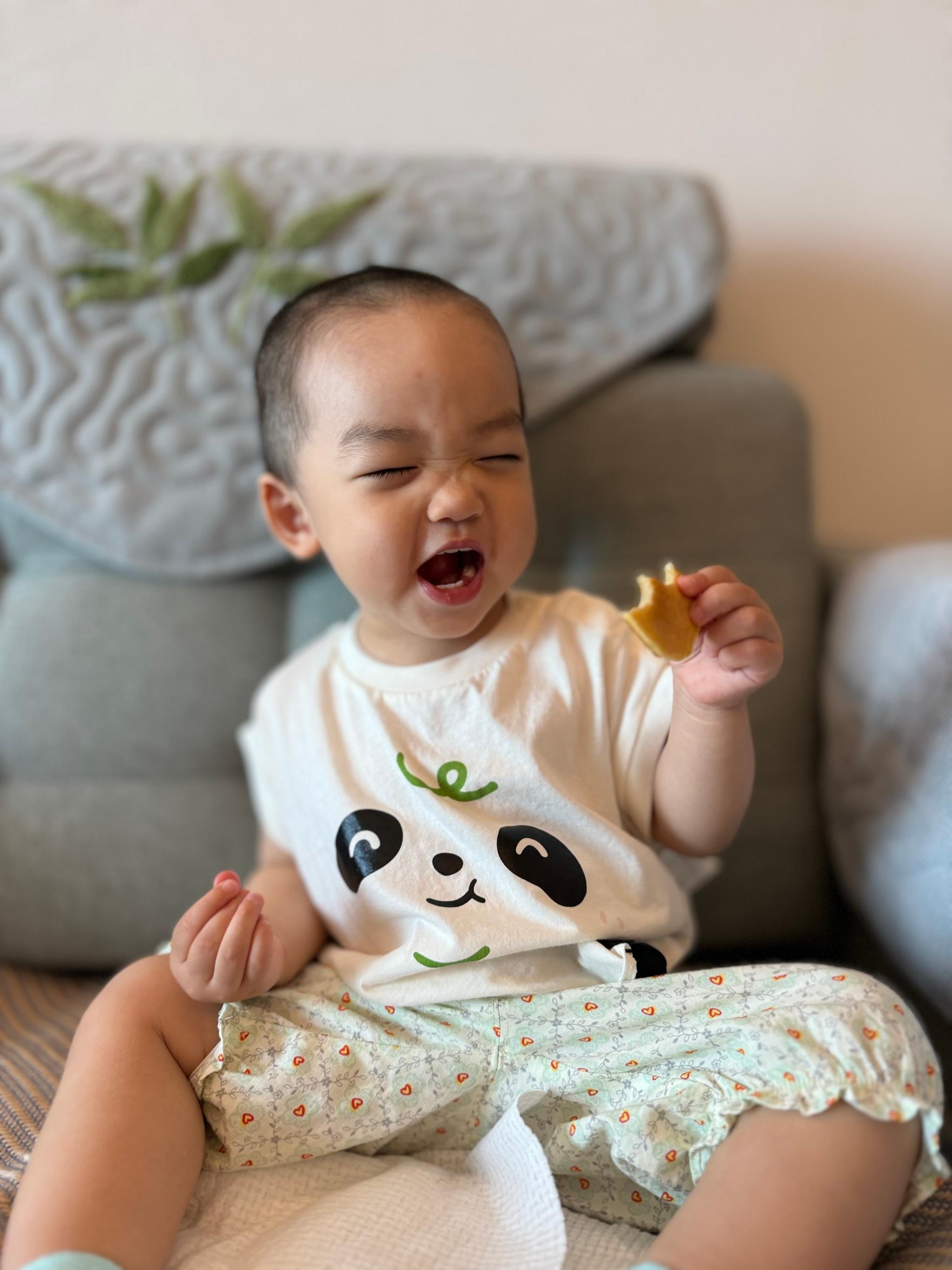 A laughing toddler with a piece of food in hand, wearing a panda-themed outfit, sitting on a couch