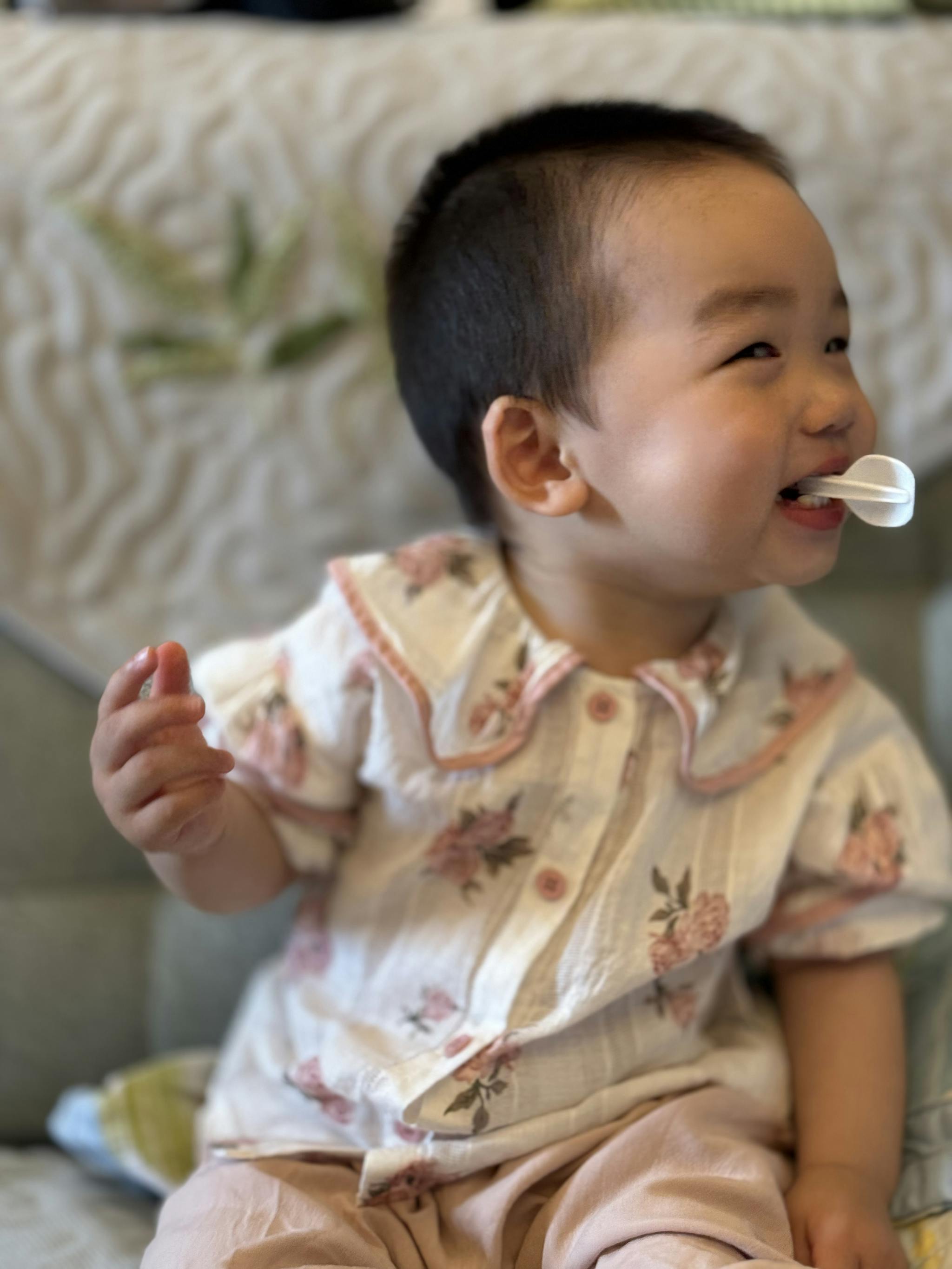 A smiling toddler in a patterned outfit sits eating from a spoon, looking joyful