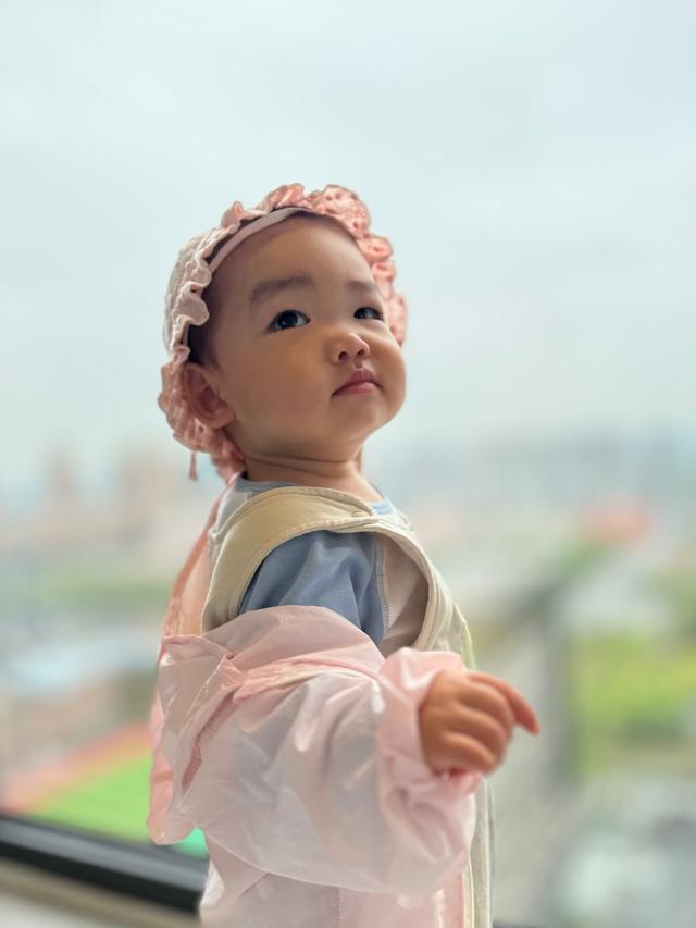 A toddler wearing a pink dress and a floral headband looks away from the camera, with a blurred background that suggests a high vantage point