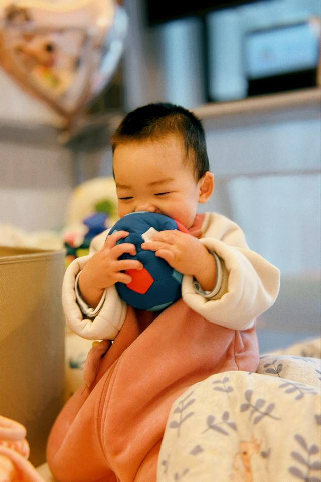A baby with a joyful expression is biting a blue ball while wrapped in a pink blanket