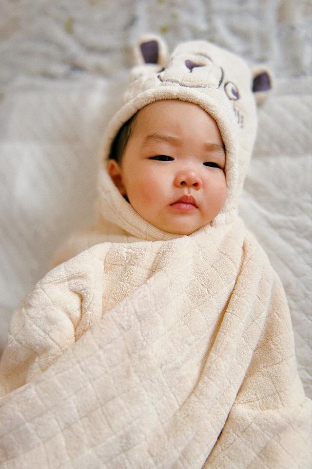A baby wrapped in a soft, cream-colored blanket with a cute animal-eared hat looks sleepily at the camera