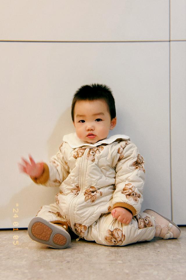 A baby sitting on the floor wearing a patterned outfit with a slightly puzzled expression, one hand raised