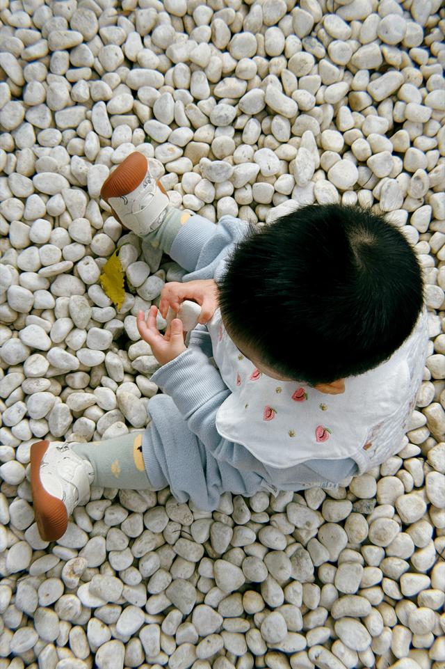 A small child is sitting on a pebbled surface, holding a half-eaten bread roll in one hand. The child is wearing a hat and a blue outfit with light-colored shoes