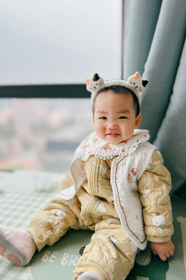 A smiling baby dressed in a yellow outfit with cute animal ears on the hood, sitting against a window