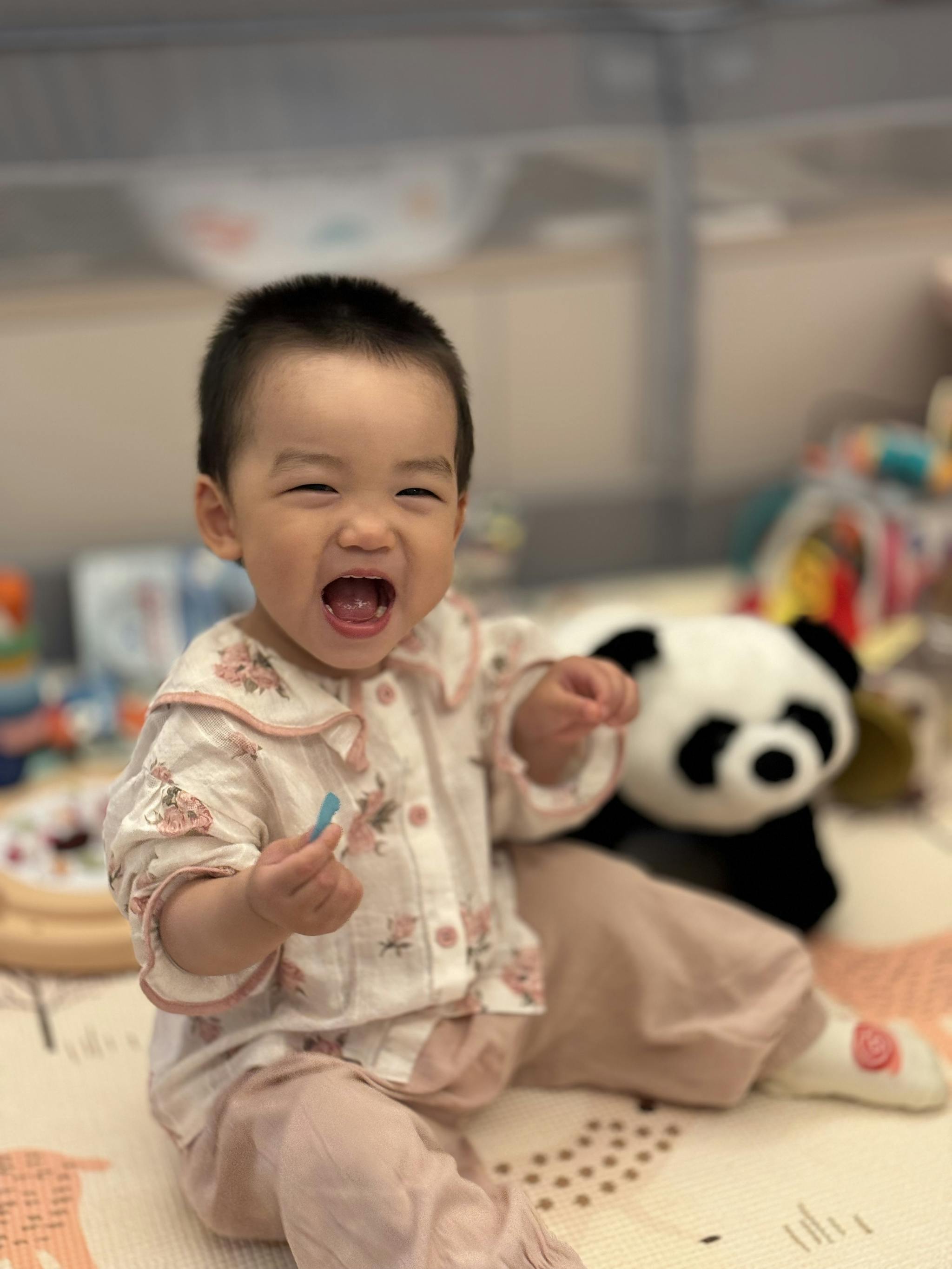 A smiling baby sits on the floor with a plush panda toy beside them, surrounded by various other toys, exuding joy and playfulness