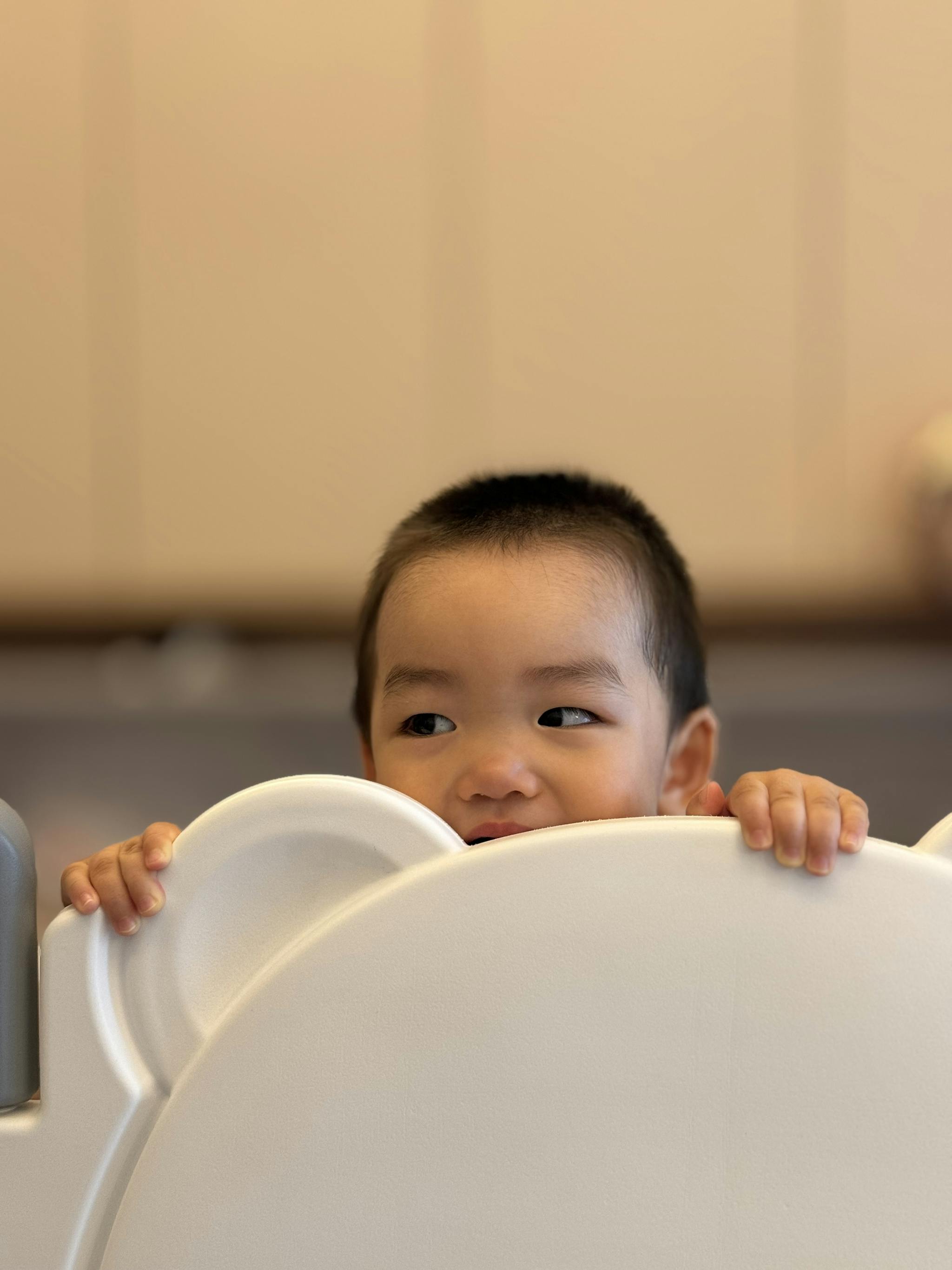 A young child peeks over the top of a white chair, only their head visible