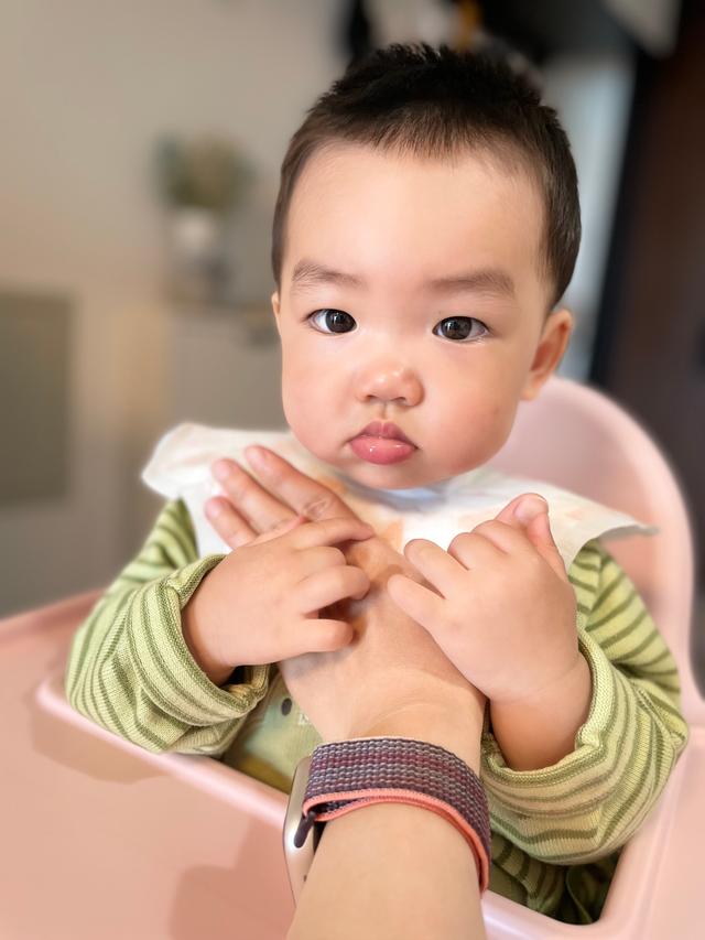A toddler with a pensive expression, wearing a striped green shirt and a bib, sits in a high chair, hands crossed in front