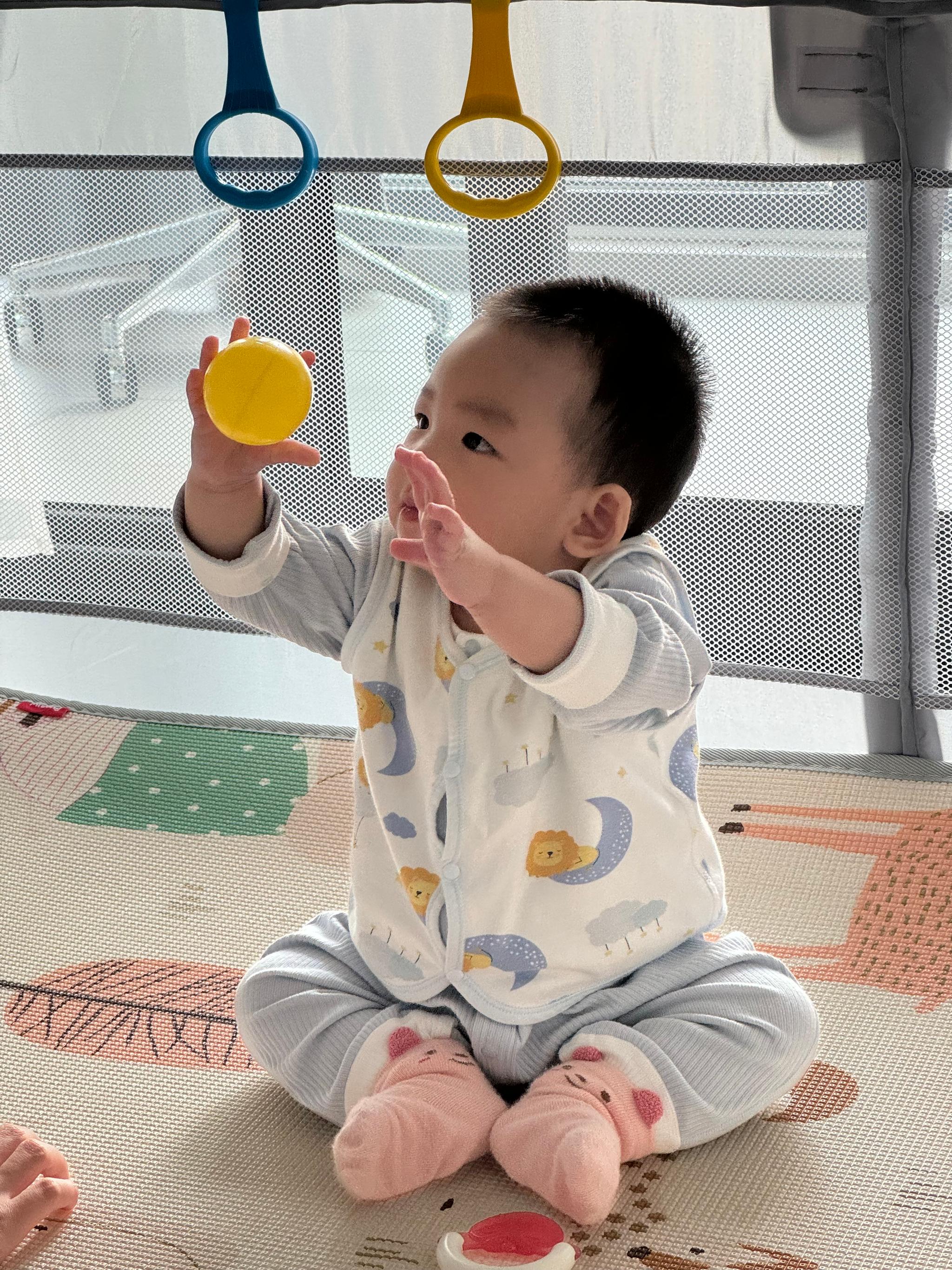 A baby sits on a patterned mat, looking curiously at a yellow toy in their hand, with other toys hanging above. The child is wearing a onesie adorned with clouds and pink sock shoes