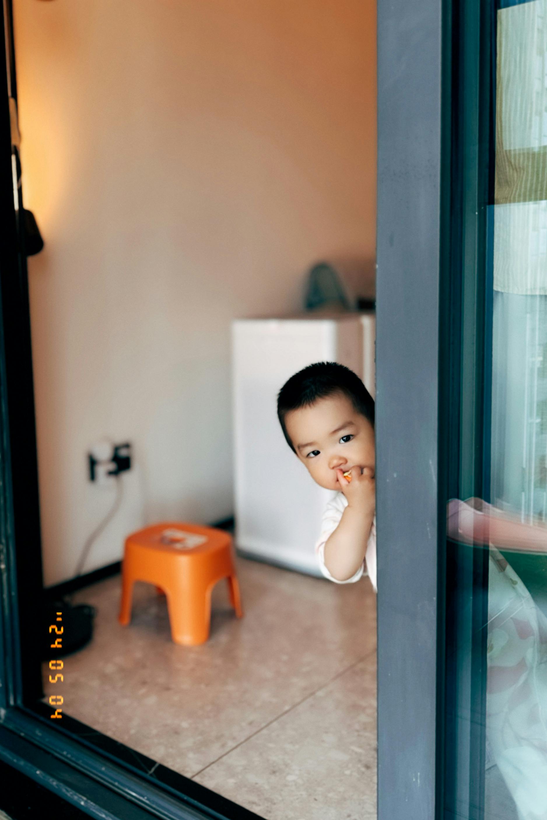 A curious child peeking out from behind a door, resting their head on their hand, with a small orange stool and a white refrigerator in the background