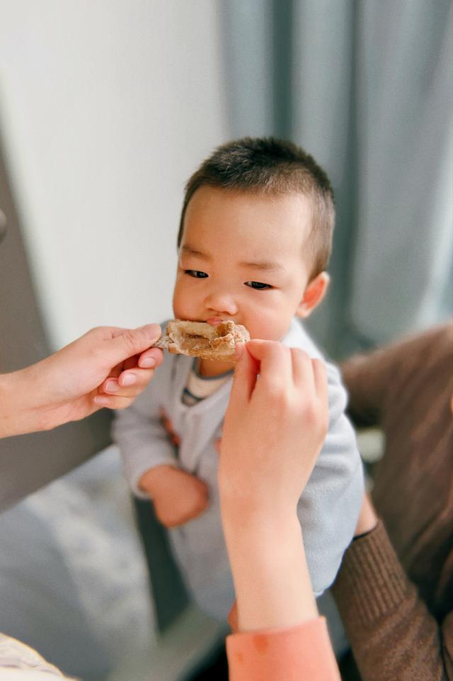 A toddler is being fed with a spoon by an adult's hand, with a slight smile on the child's face indicating enjoyment or contentment