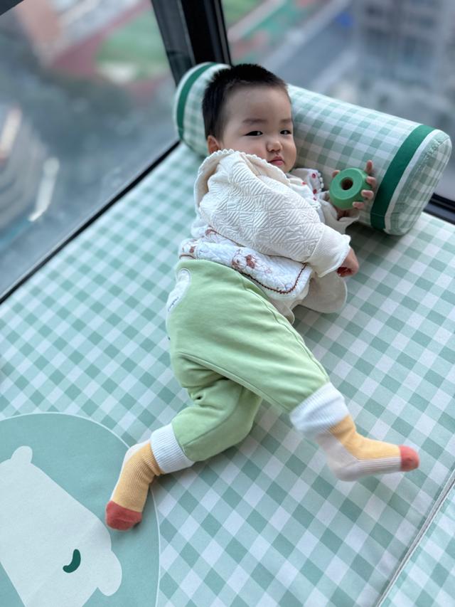 A baby with a playful expression is lying on their stomach, propped up on their arms, wearing a white blouse and green pants with red socks. There's a green toy nearby, and the setting appears to be indoors with natural light coming in from the left