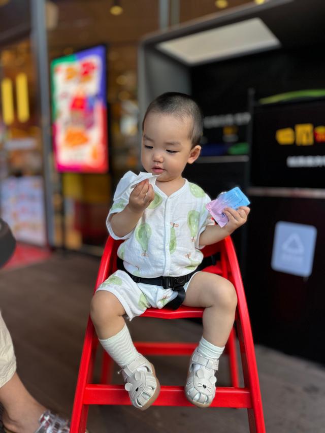 A young child sitting on a red chair, holding a piece of food in one hand and a colorful object in the other, with a background of a storefront and bright signage