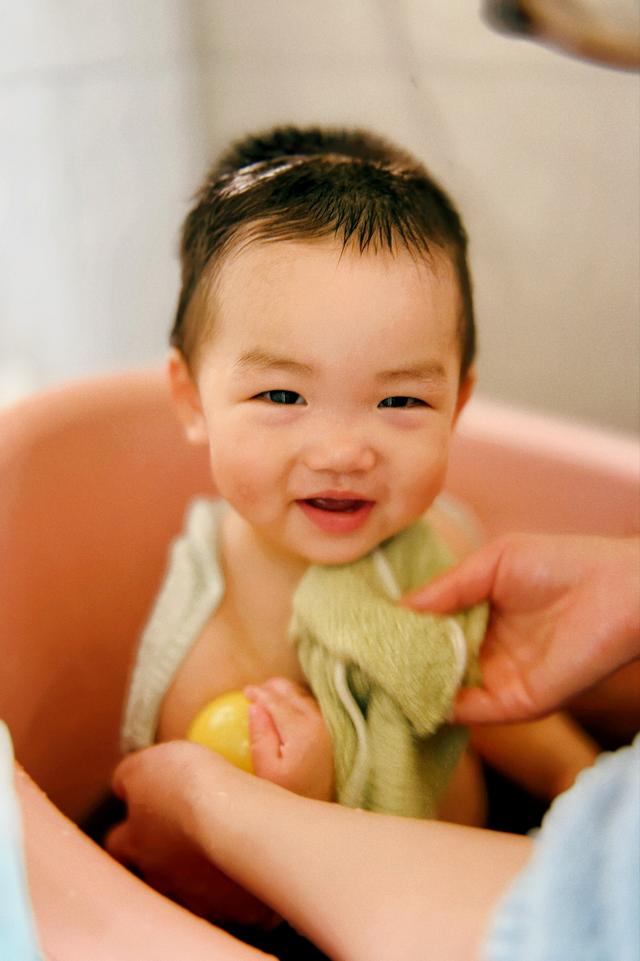 A smiling baby wrapped in a yellow towel is sitting in a pink basin, likely during or after bath time