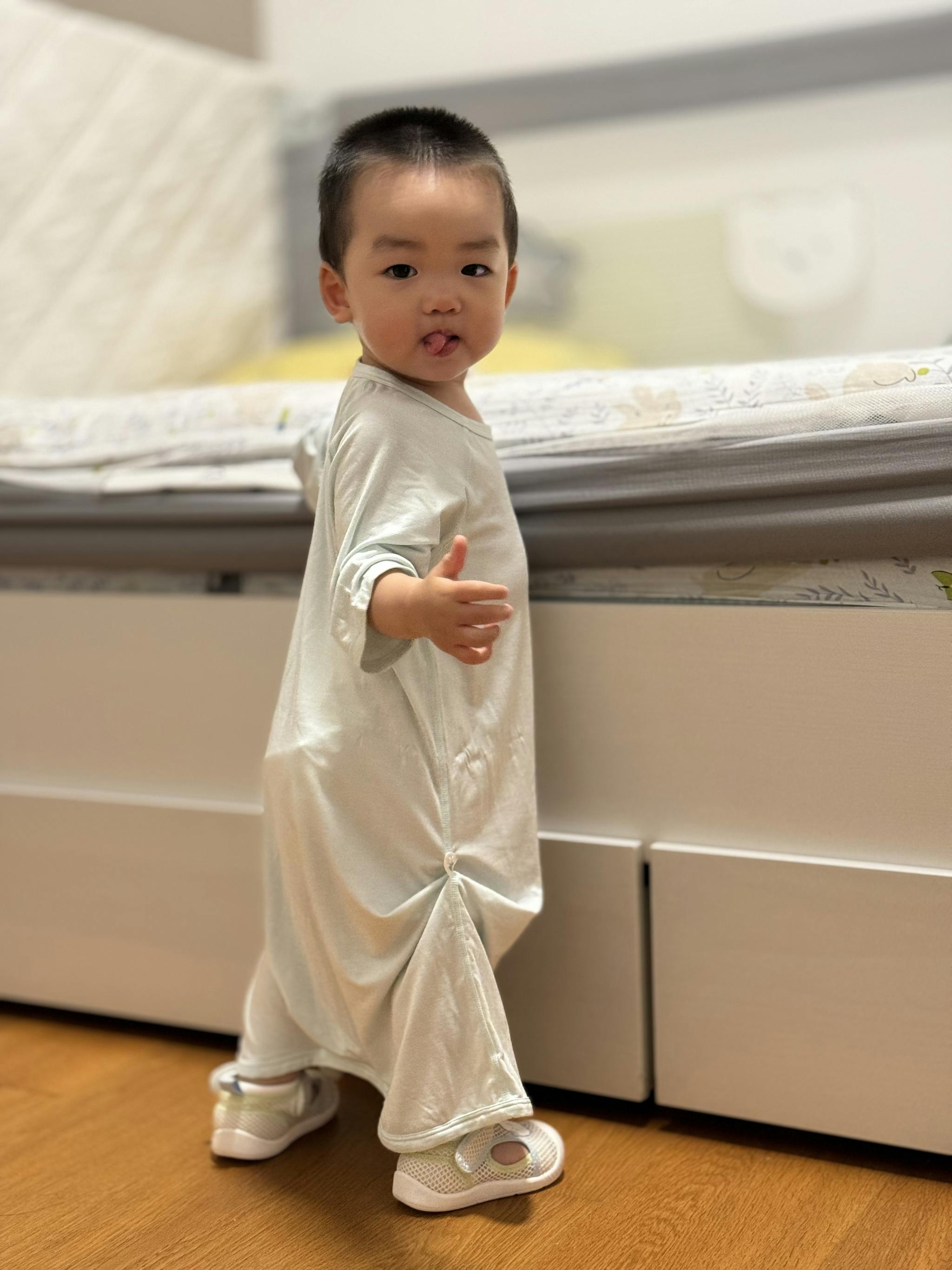 A toddler standing indoors wearing a long white garment with a curious expression, looking over their shoulder