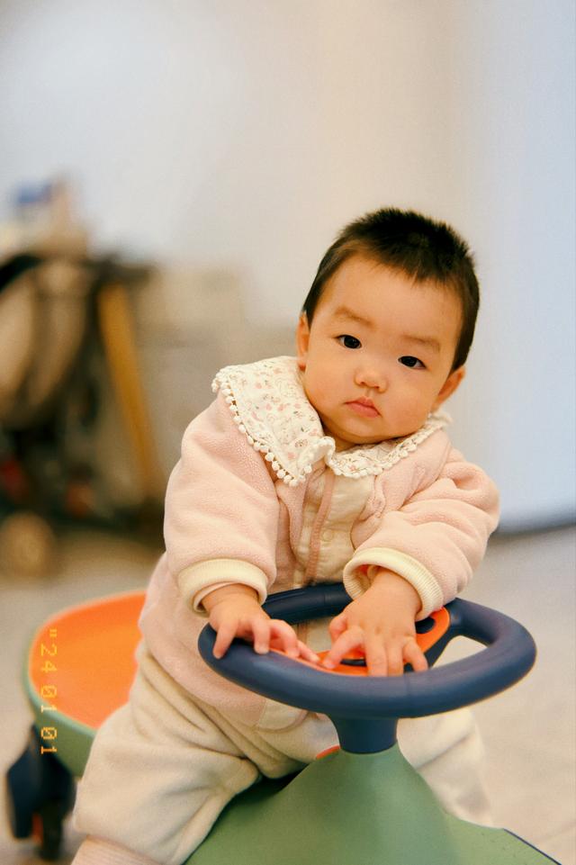 A baby with a neutral expression sits grasping the steering wheel of a toy car, dressed in light-colored clothing with a blurred background suggesting indoor surroundings