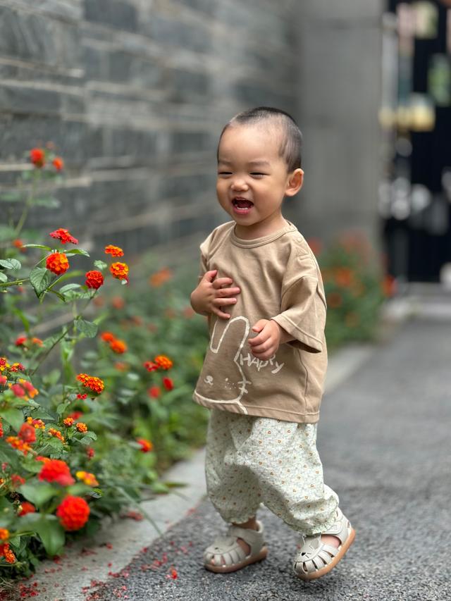 A toddler stands smiling beside orange flowers, wearing a beige top and patterned pants, with a blurred background emphasizing the child