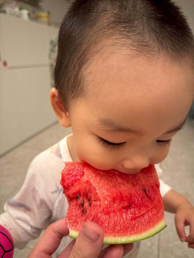 A young child is eating a large slice of watermelon, with an adult's hand holding the fruit