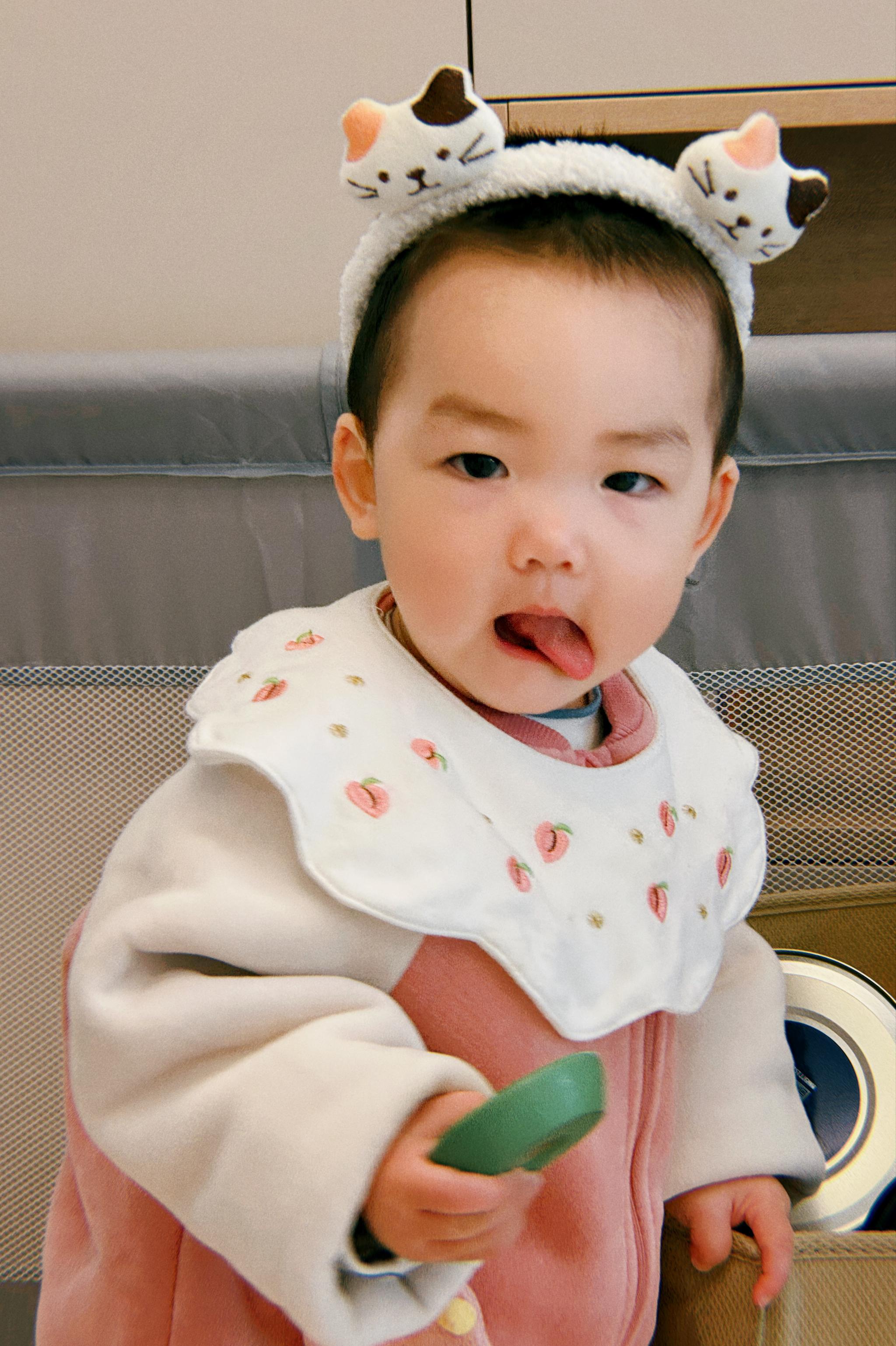 A toddler with a whimsical expression, wearing a white bib and a headband with decorative ears, sticks out their tongue