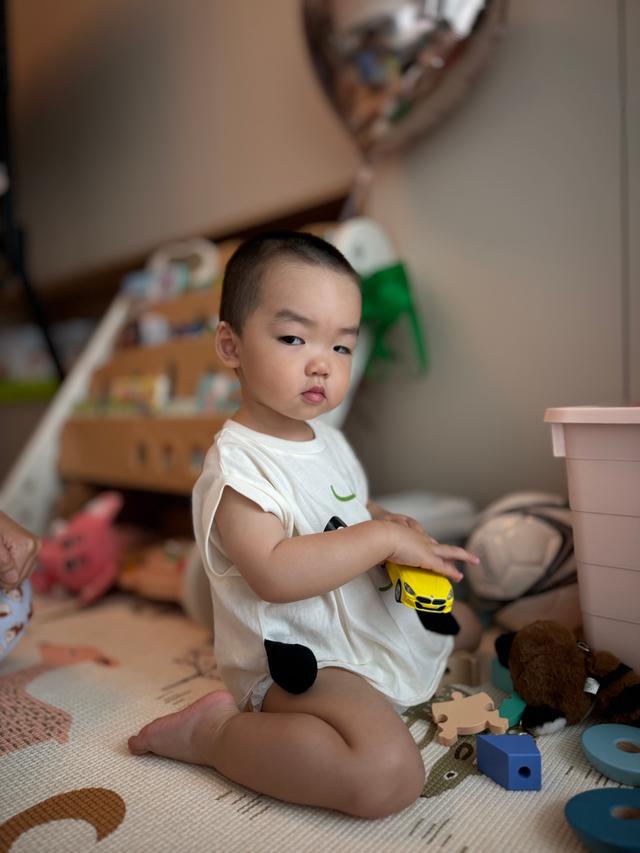 A toddler sits on the floor surrounded by toys, holding a yellow toy car, with a focused expression on their face