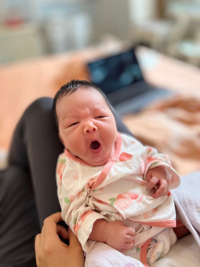 A yawning baby wrapped in a floral blanket, sitting on someone's lap