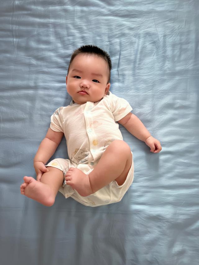 A baby with a playful expression is lying on a blue background, dressed in a cream outfit and looking directly at the camera