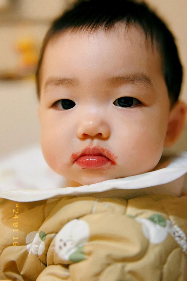 A baby with a serious facial expression, wearing a bib and patterned clothing, appears to have just eaten, as suggested by the reddish residue around the mouth